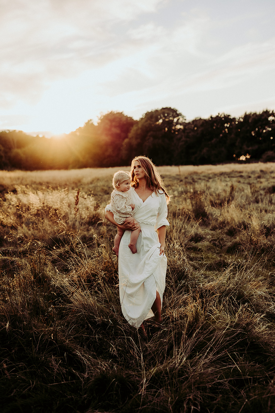 en mamma i en bohemisk vit klänning bär sitt barn när hon går över ett fält