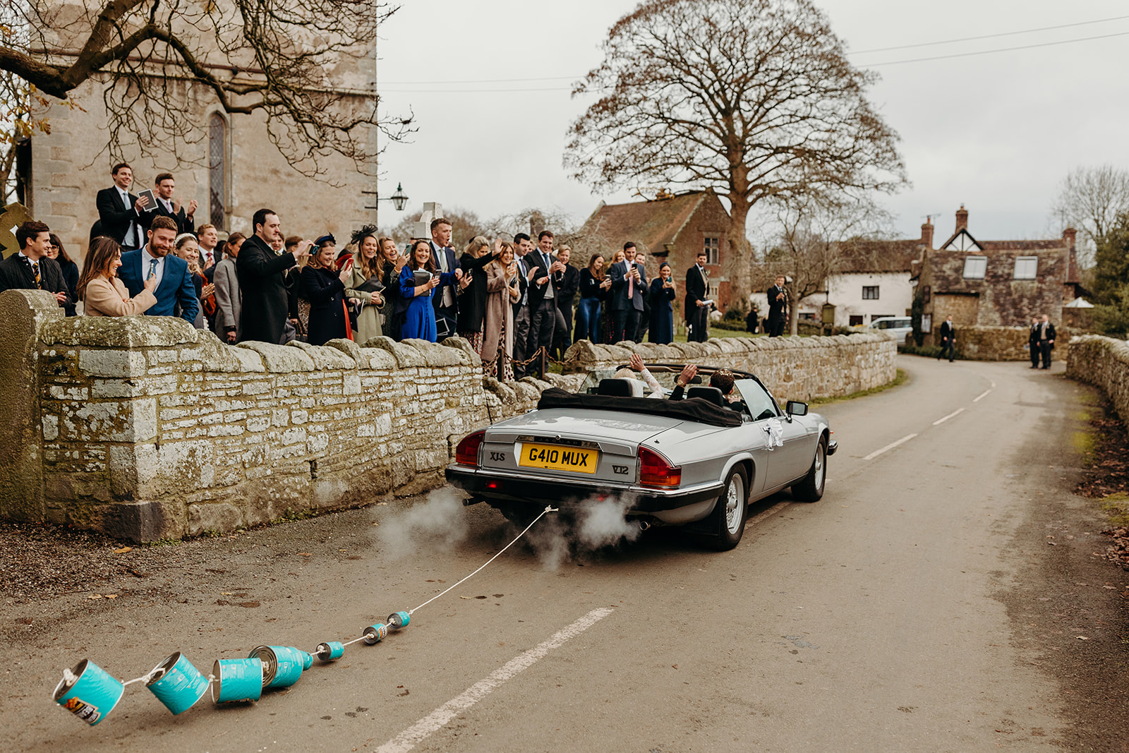 bride and groom leaving in vintage car with cans tailing behind as guests wave them on