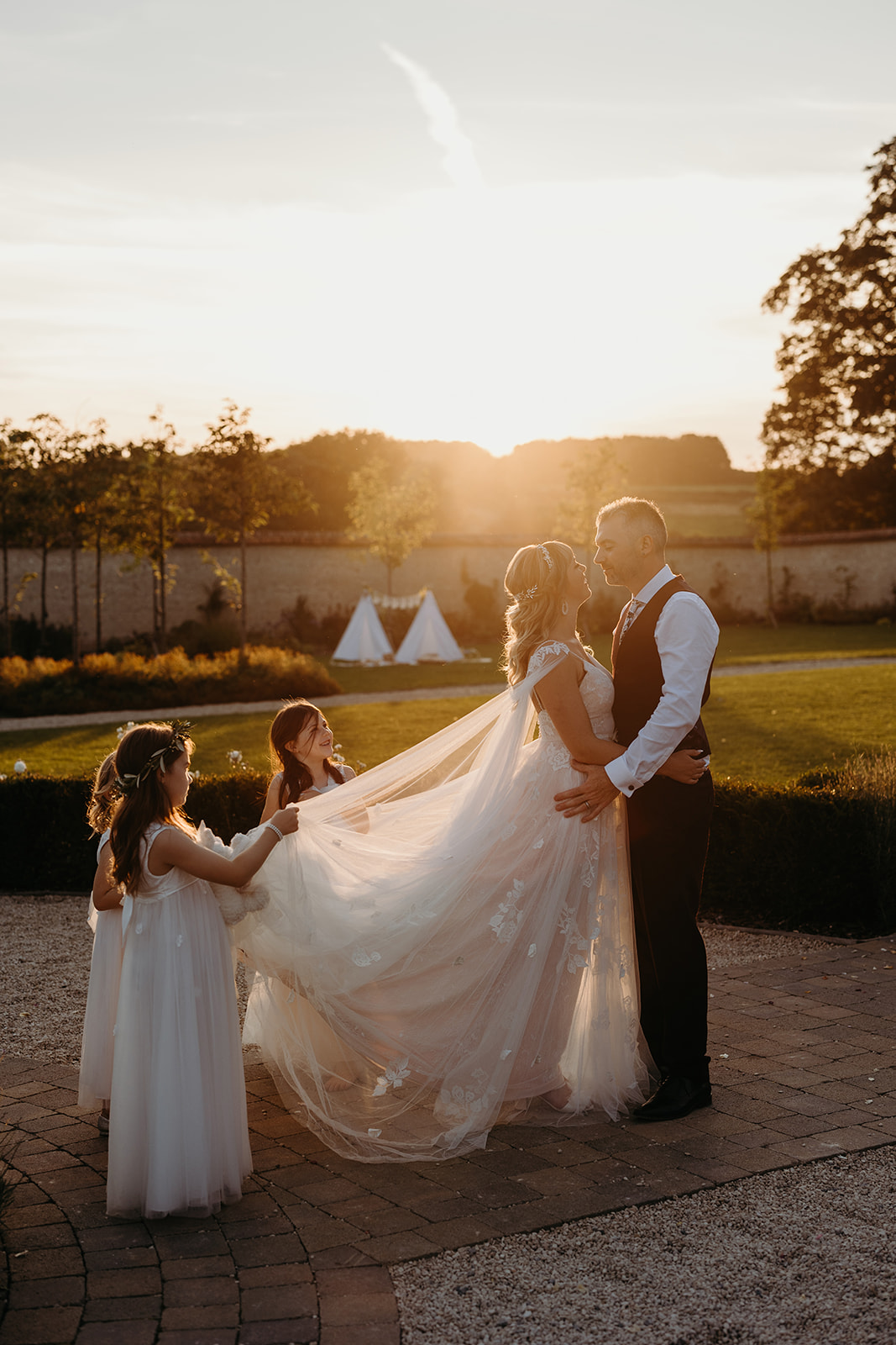 sunset flow as flower girls help bride with dress as she stands with groom