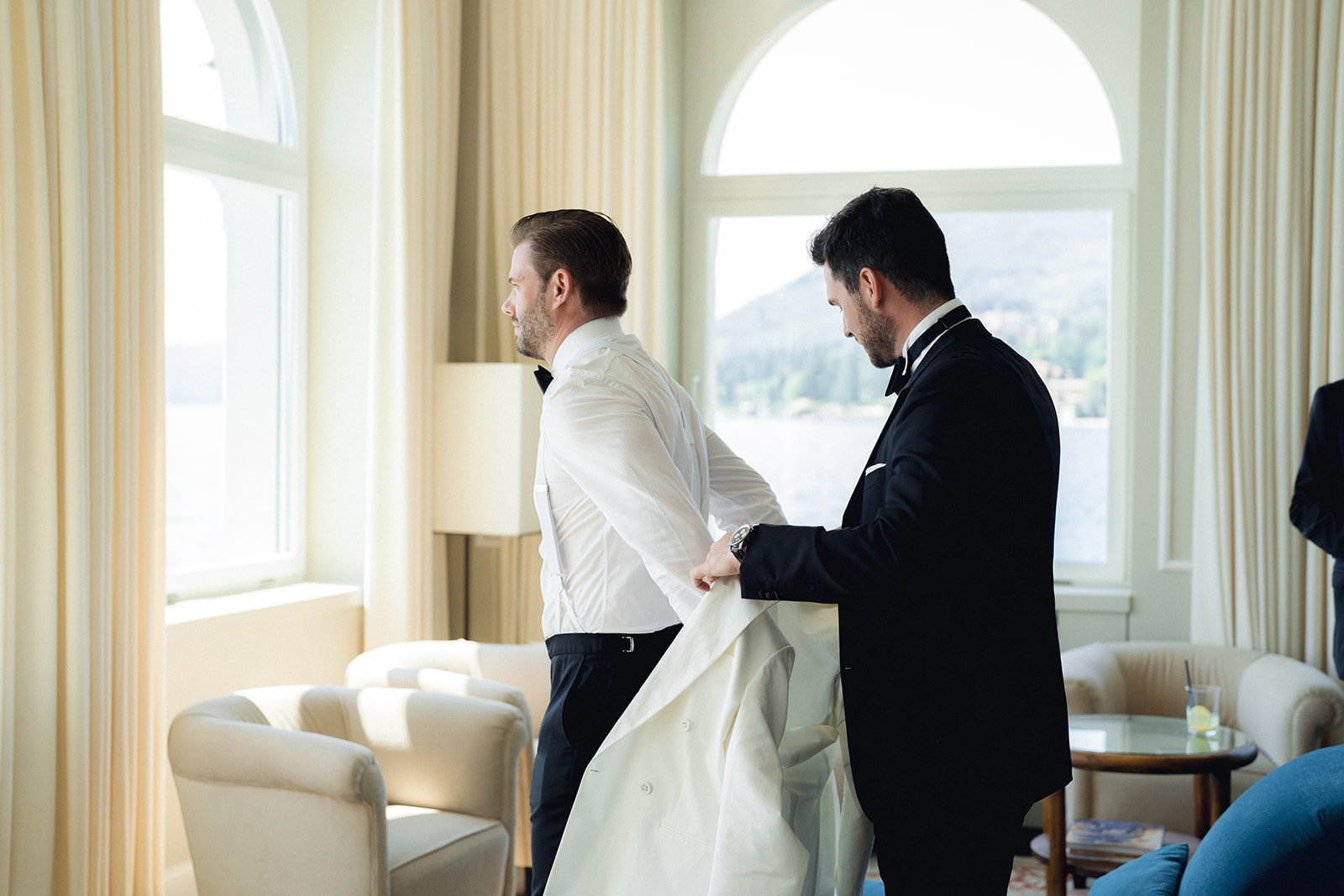 The groomsman helps the groom put on the white jacket of the tuxedo for his wedding