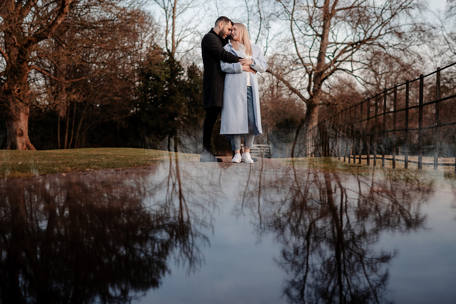 A couple stand with his arms wrapped around her on a country path, the treeline reflected on the ground at their feet