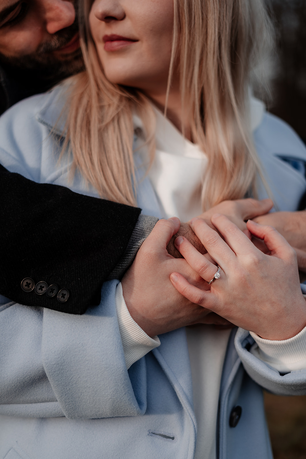 A couple's hands clasped together as he wraps his arms around her, her engagement ring clearly visible
