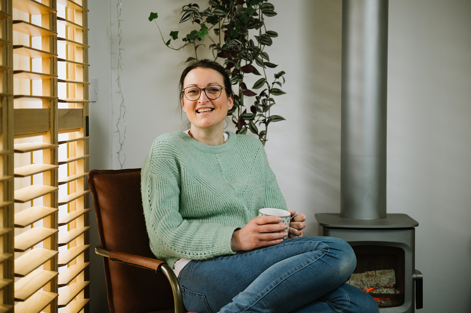 smiling woman holding cup of tea wearing mint green jumper with plants and a log burner in the background