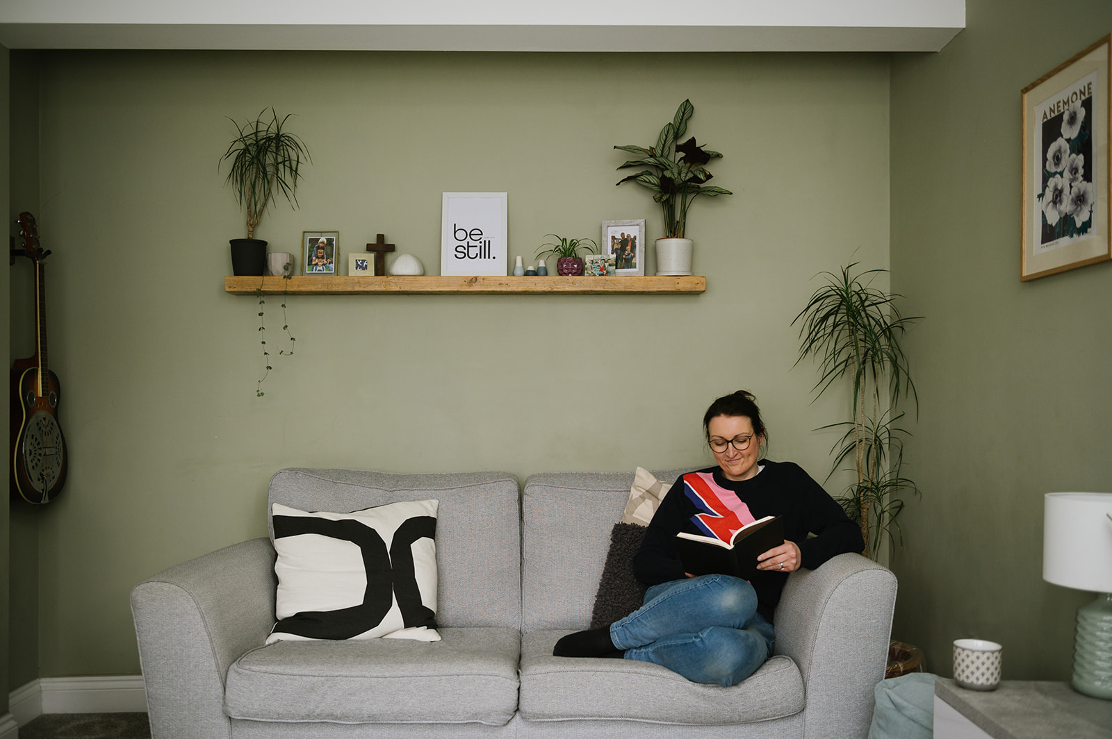 a woman smiling reading a book on the sofa in a long with a shelf of decorative objects above her