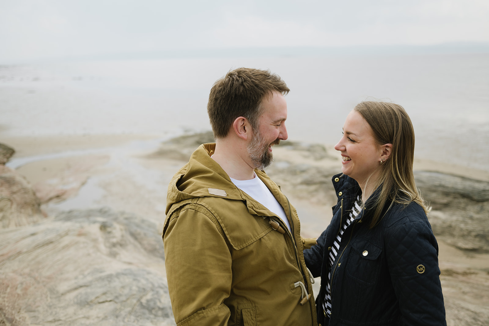 couple facing each other smiling, they're wearing coats with a beach blurred in the background