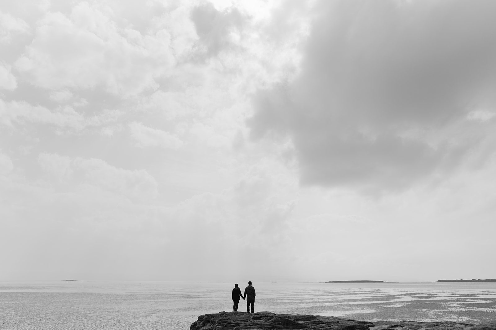 A couple in silhouette in the distance stand holding hands faceing away from the camera looking out to sea