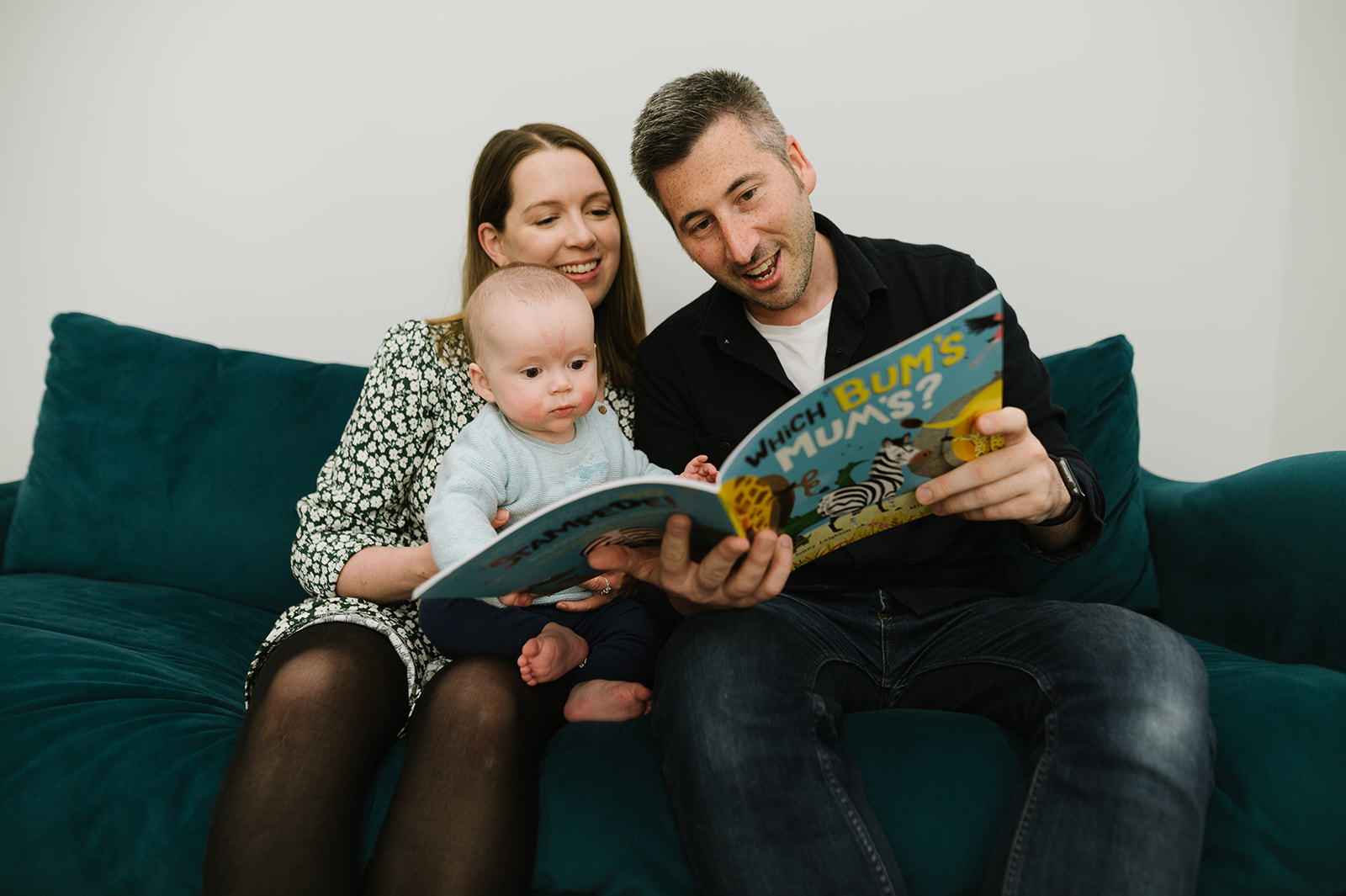 mum, dad and baby reading a book together on a blue sofa 