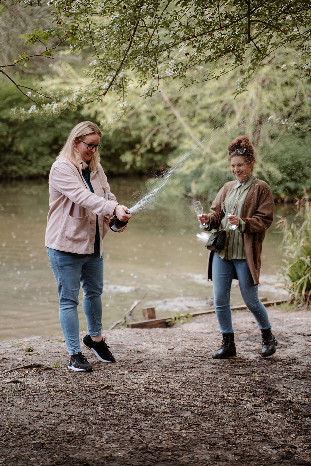 female couple pop a bottle of prosecco while stood by a lake - couples photography in Hampshire