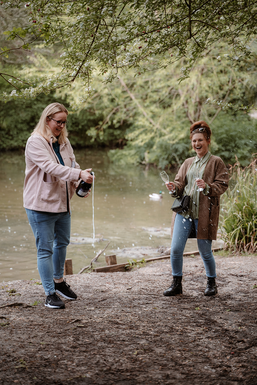 female couple pop a bottle of prosecco while stood by a lake - couples photography in Hampshire
