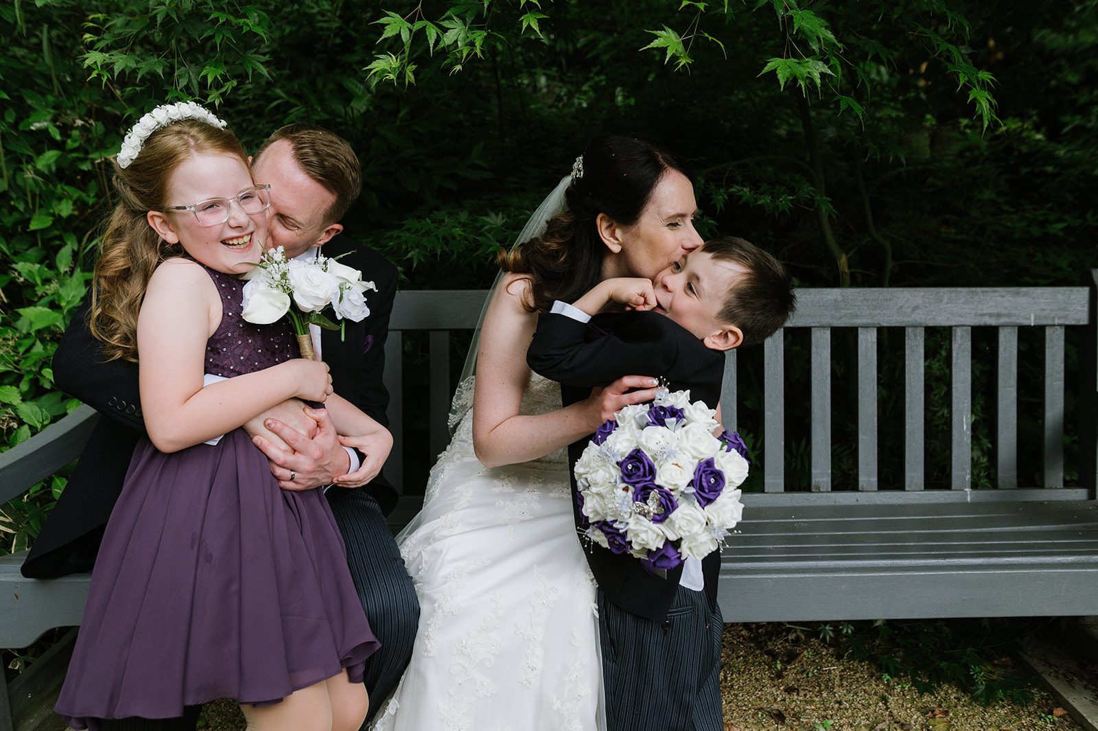 bride and groom with their young children sitting on a bench kissing the kids on the cheek