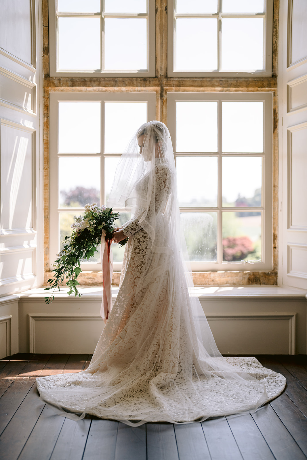 bride with veil over her head in lace wedding dress in front of window backlit