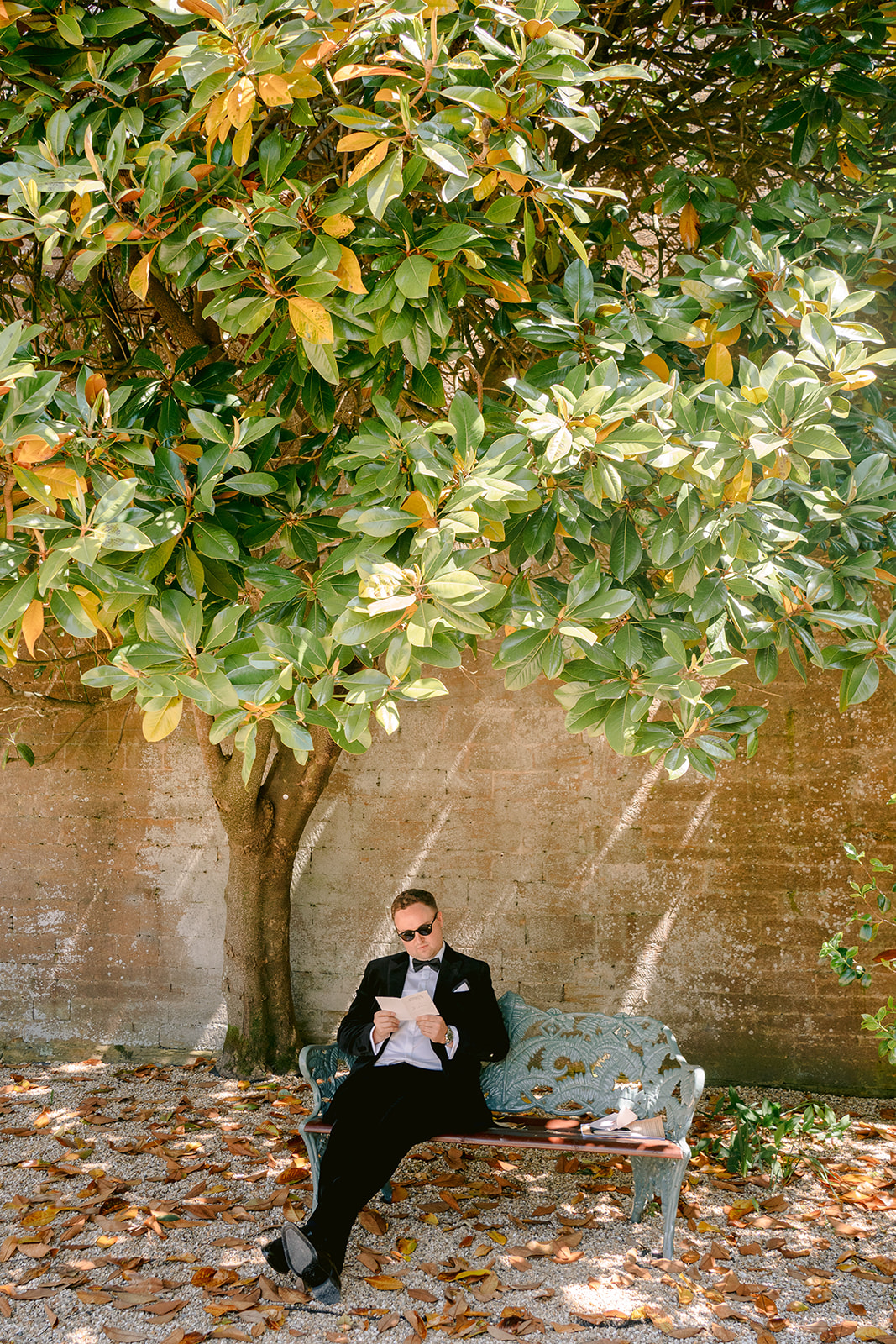 groom reading his wedding card on the bench under a tree outside brympton houuse