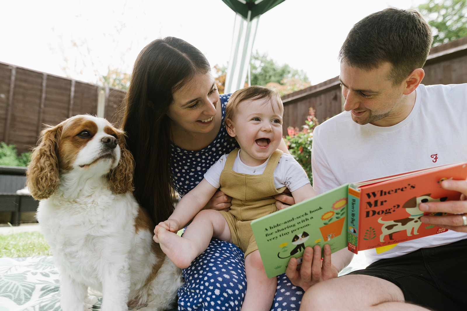 mum dad and one year old boy smiling as they read their book in the garden. their dog is sitting with them