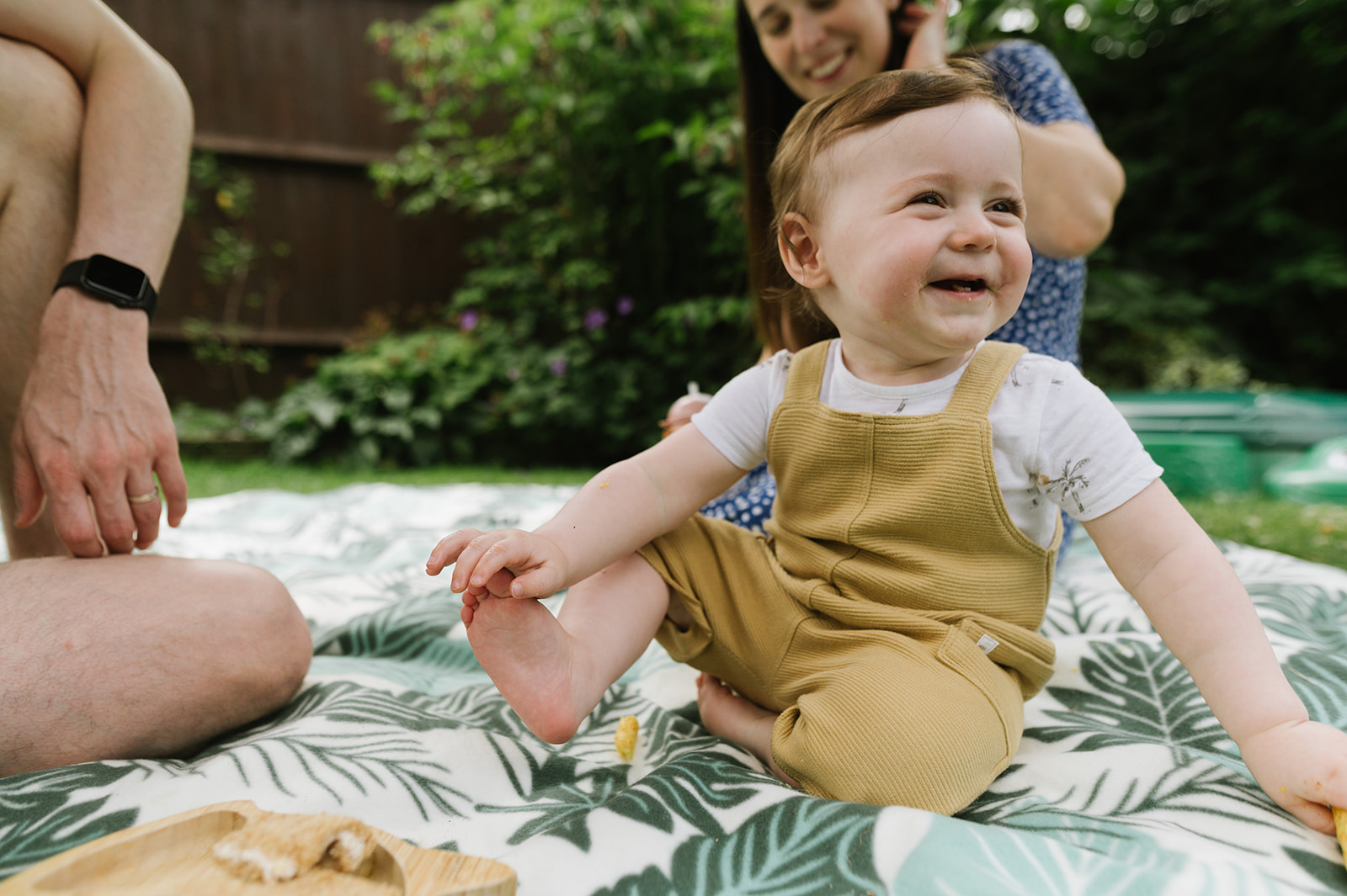 one year old baby boy smiling sitting on a picnic rug in the garden holding his foot 