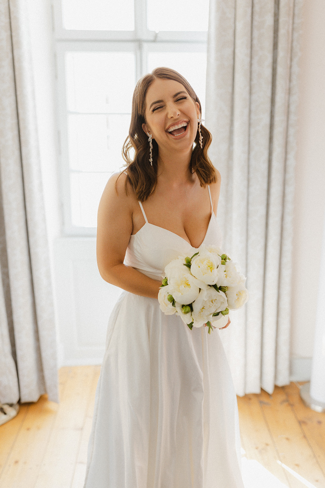 Elegante Braut steht vor dem Fenster im Weingut Boudier Koeller und hält einen Strauß aus weißen Pfingsrosen in der Hand