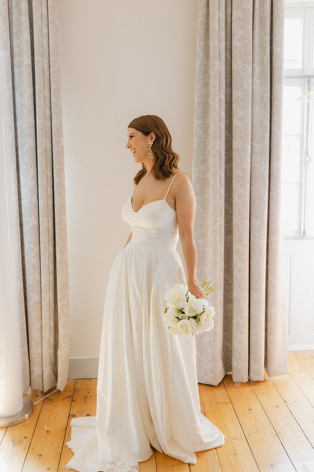 Elegante Braut steht vor dem Fenster im Weingut Boudier Koeller und hält einen Strauß aus weißen Pfingsrosen in der Hand