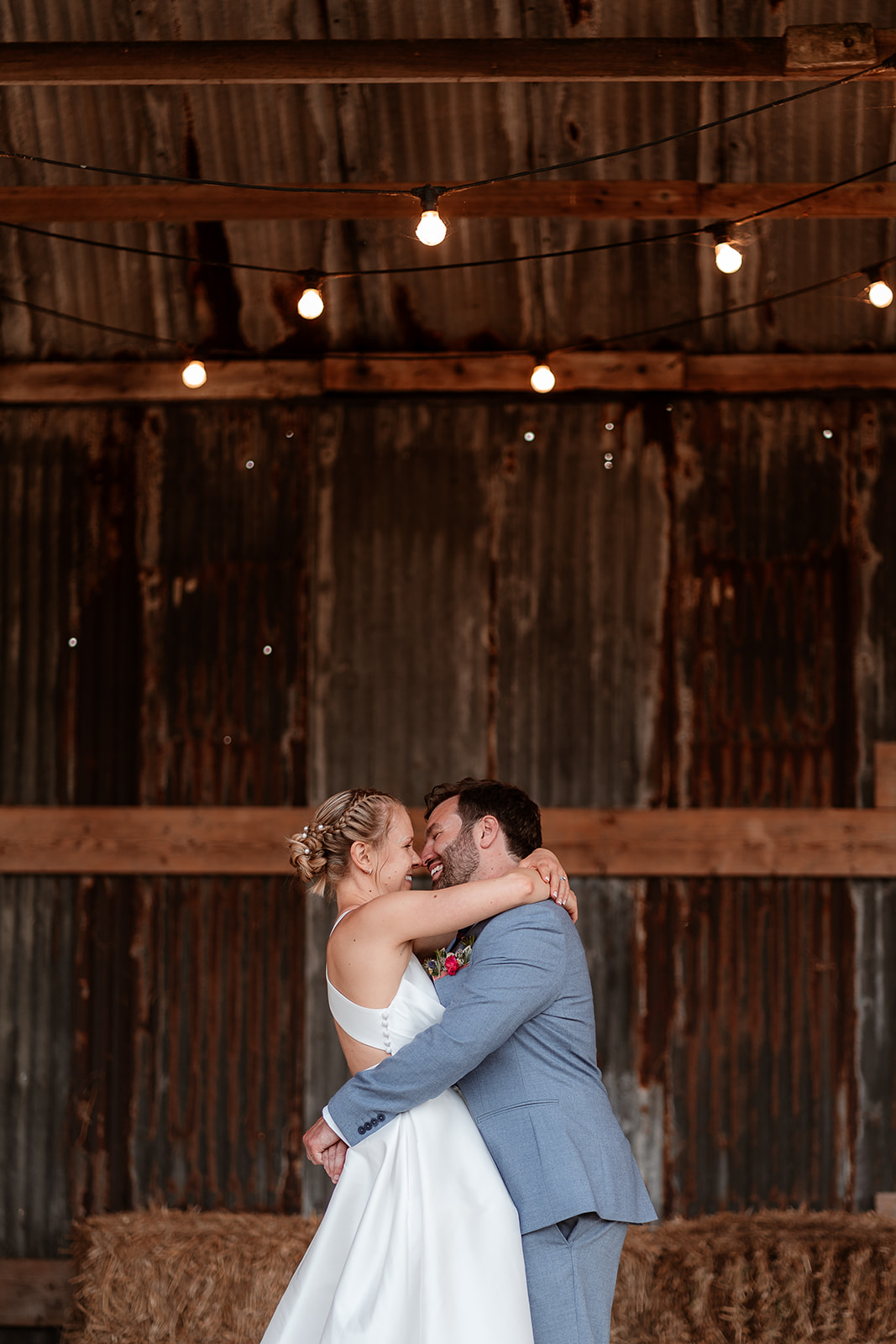 Bride and groom cuddle under the festoon lights in the Dutch Barn at a summer wedding at Silchester Farm