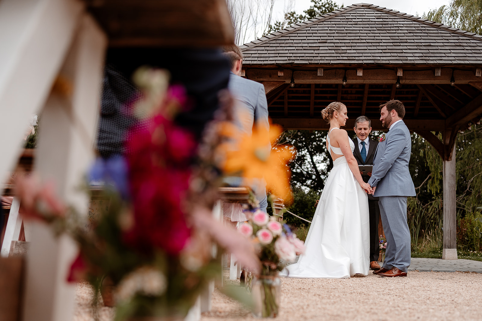 Bride and groom hold hands during their ceremony with bright summery flowers in the foreground - Silchester Farm wedding