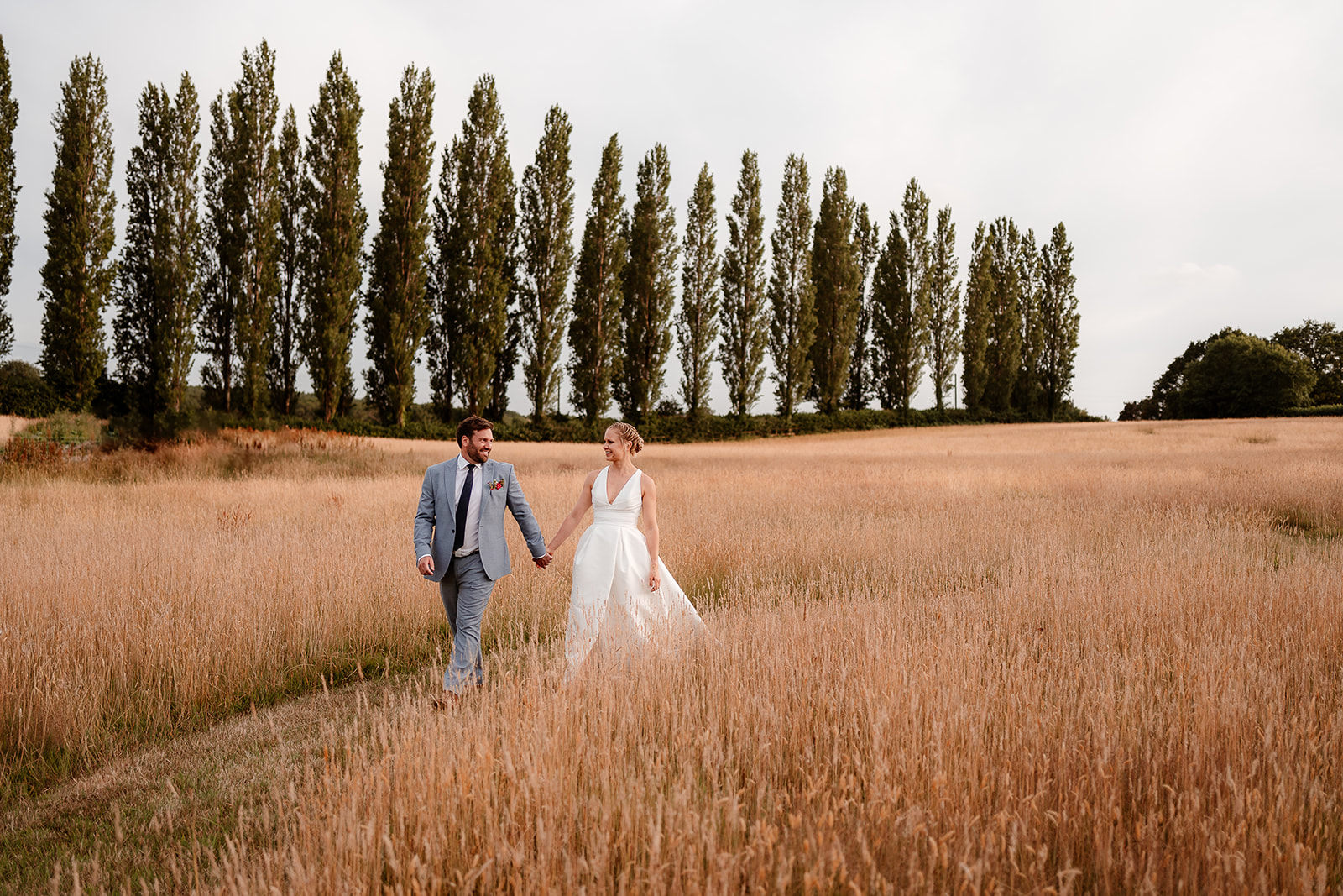 Bride and groom walk hand in hand through the long grass at sunset at a summer wedding at Silchester Farm