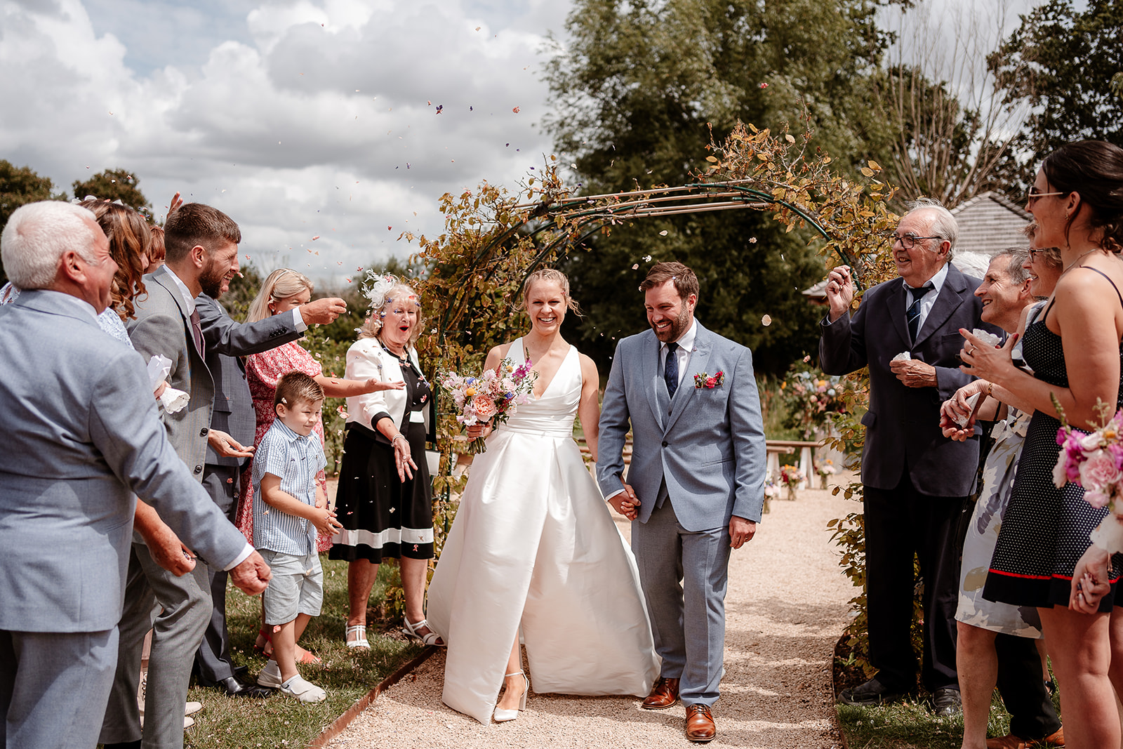 Bride and groom walk through a confetti tunnel, beaming with joy at a summer wedding at Silchester Farm