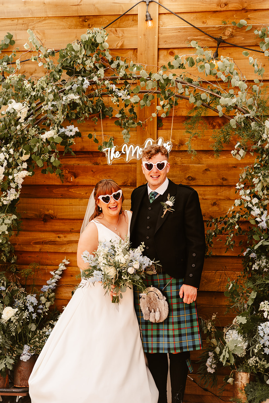 Bride and groom standing in front of a greenery arch, neon sign and rustic wooden panels at Elrick House Wedding Venue