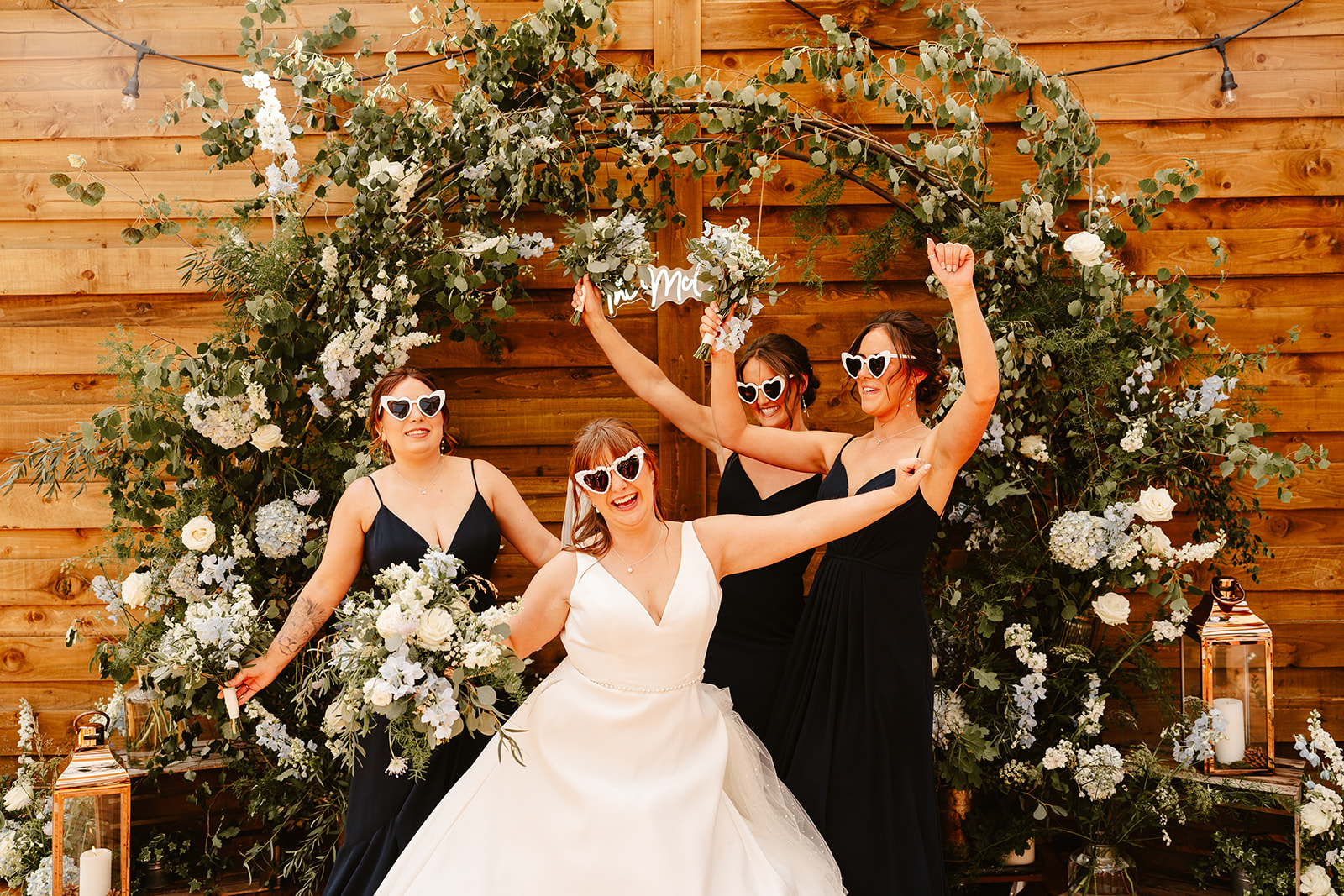 Bridesmaids wearing heart shaped sunglass standing in front of a greenery arch, neon sign at Elrick House Wedding Venue