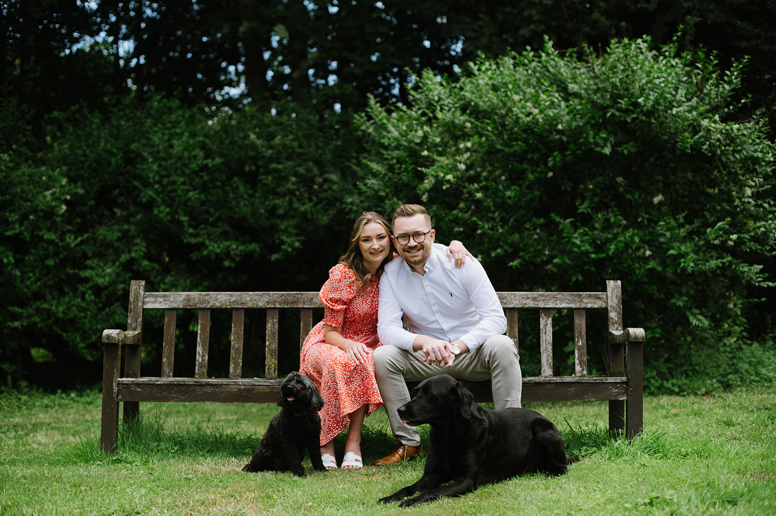 Couple seated on a bench with their 2 black dogs. Shes wearing a red dress, he's in a white shirt and chinos