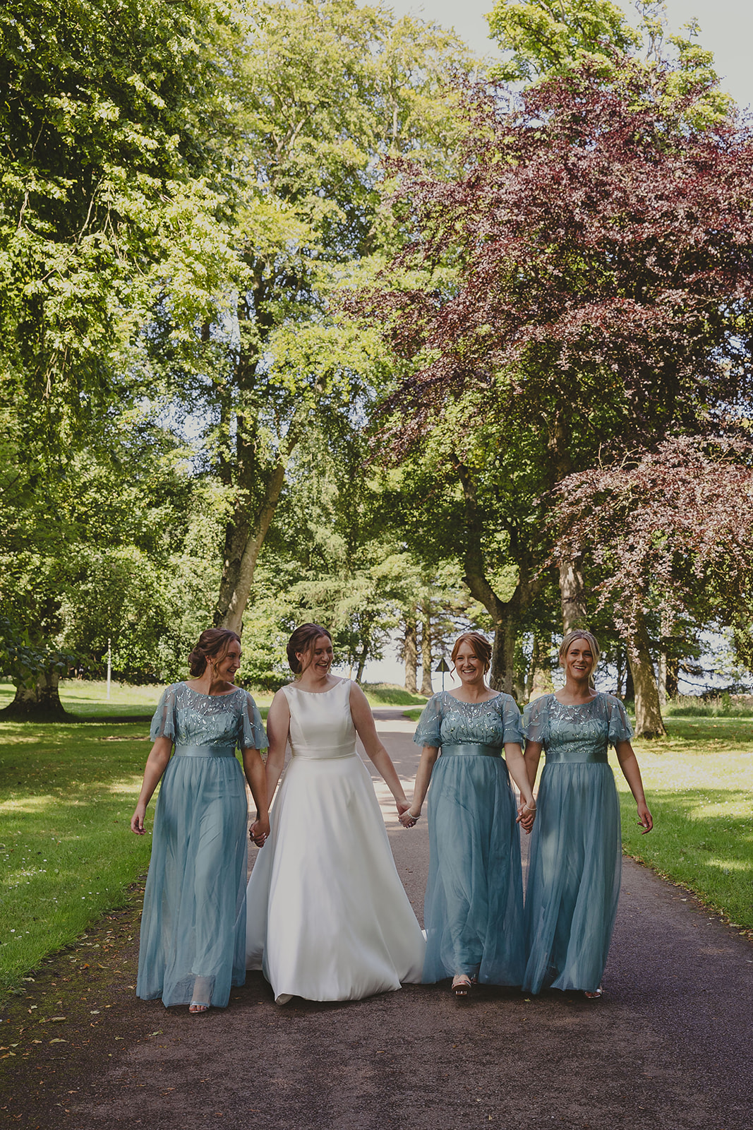 Three bridesmaids dresses in turquoise hold hands with a bride as the walk along a path at Elsick House on a wedding day