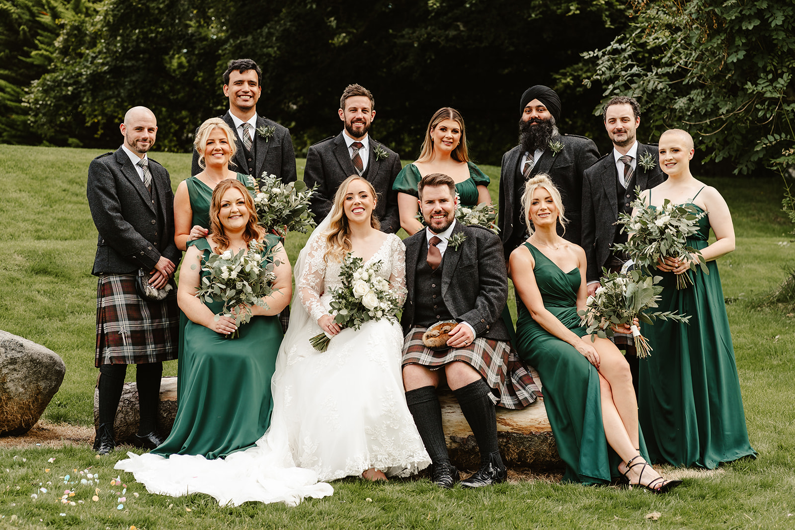 Bride and Groom sitting bridal party. Bridesmaids are wearing green dresses and groomsmen wearing kilts