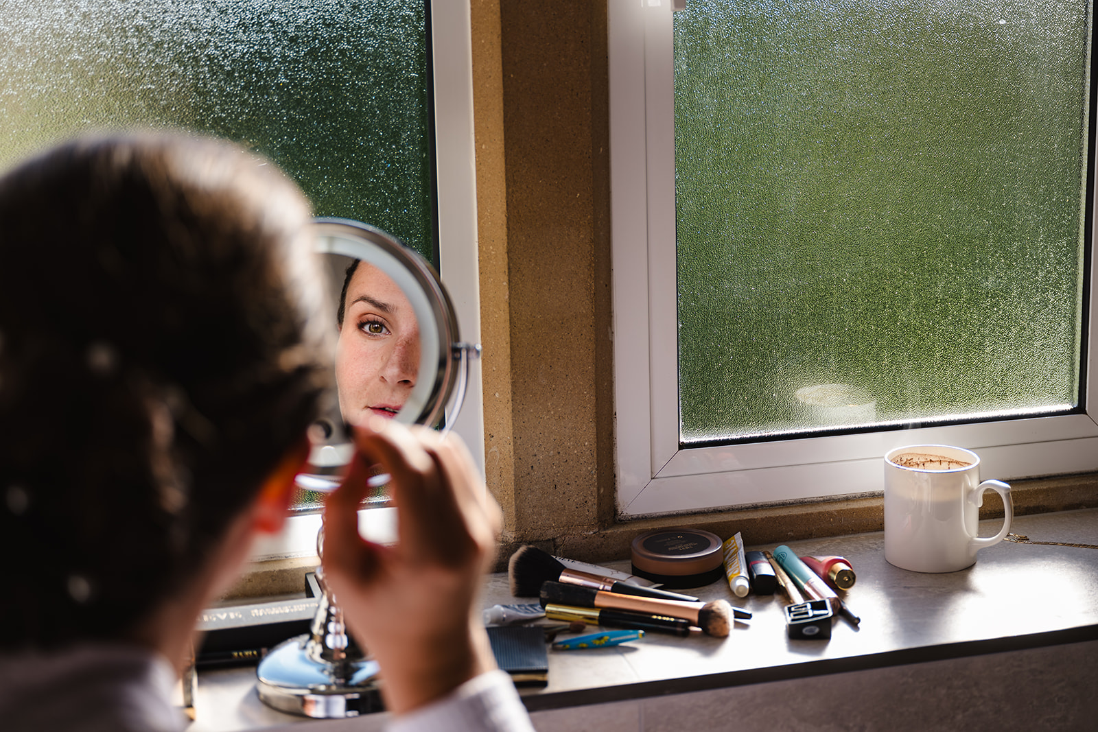 Bride applying makeup in a small mirror with a coffee steaming on the side