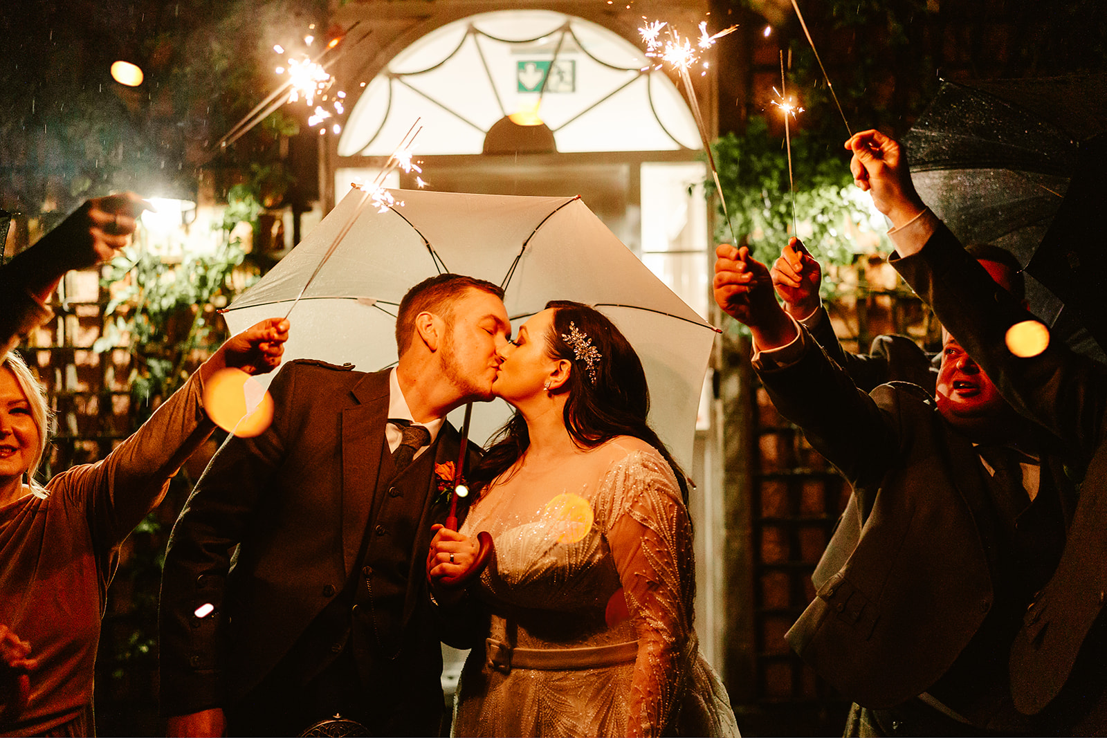 couple stand under an umbrella and kiss whilst their friends hold sparklers in the dark at ballogie house