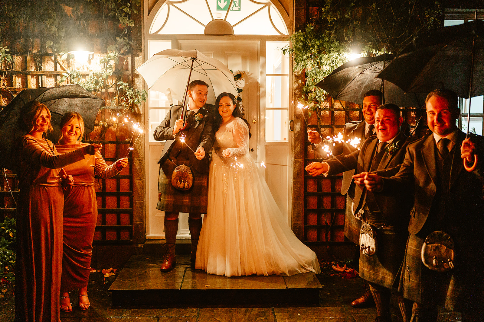 couple hold sparklers under umbrella at night time with bridal party outside Ballogie House wedding venue