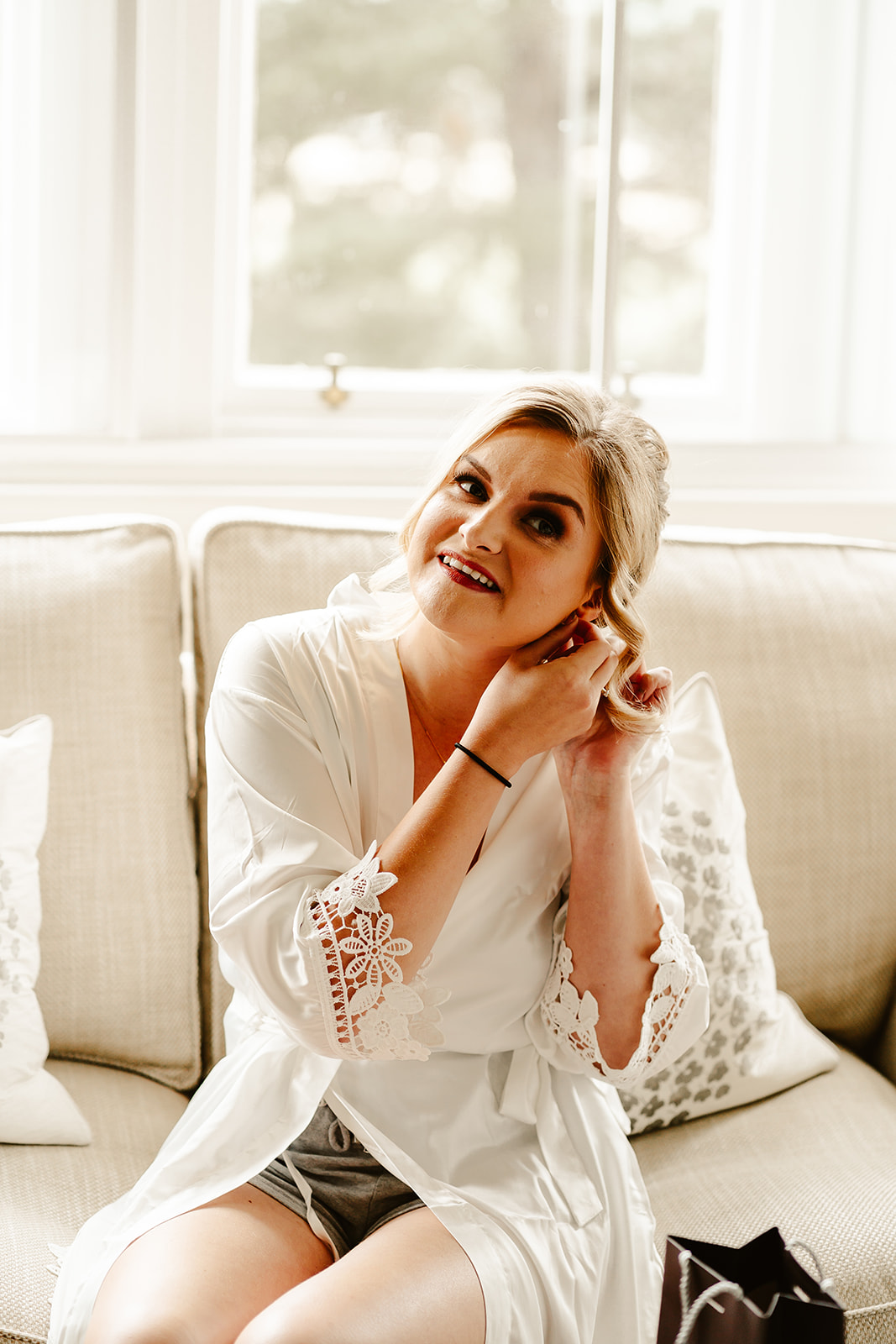 bride is putting her earrings in as she sits on a sofa in her morning robe