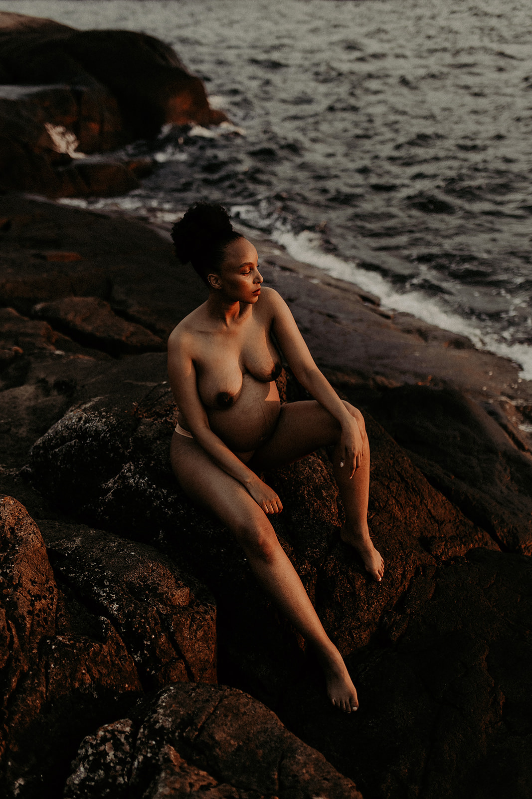En vacker, gravid kvinna sitter naken på klippor och blickar ut över solnedgången