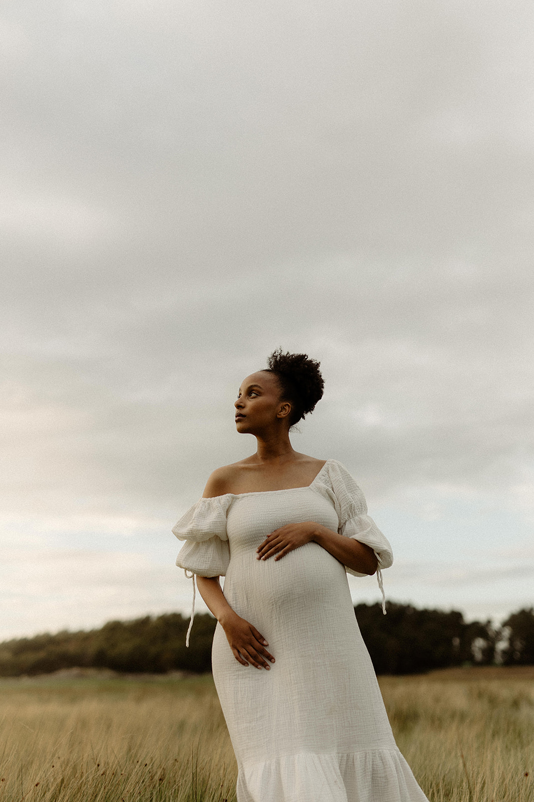 På ett fält i Göteborg står en gravid kvinna och blickar avslappnat upp mot himlen. Hon bär en vit gravidklänning.