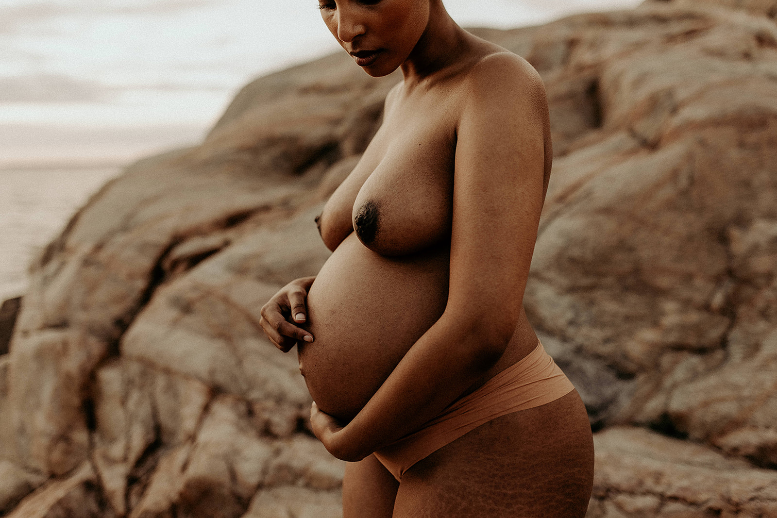 I solnedgången bland klipporna står en mamma och håller om sin gravida mage endast iklädd trosor