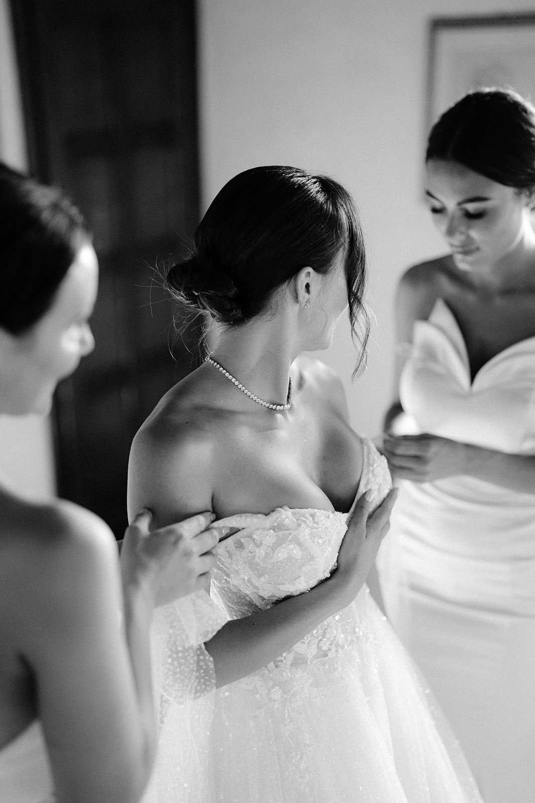 The bride is wearing a strapless gown, assisted by the bridesmaids