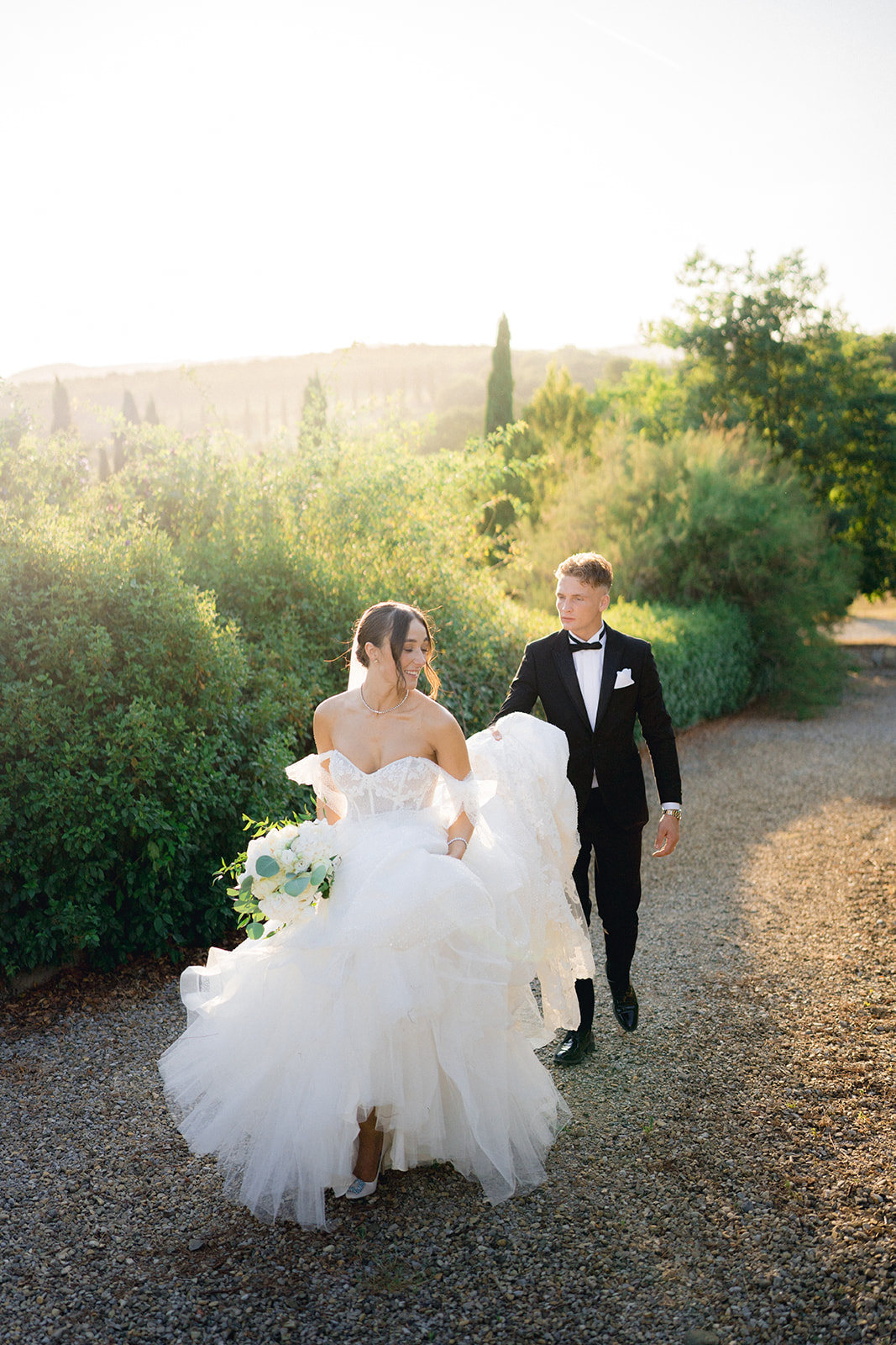 The groom helps the bride with the train of her dress as they walk on gravel