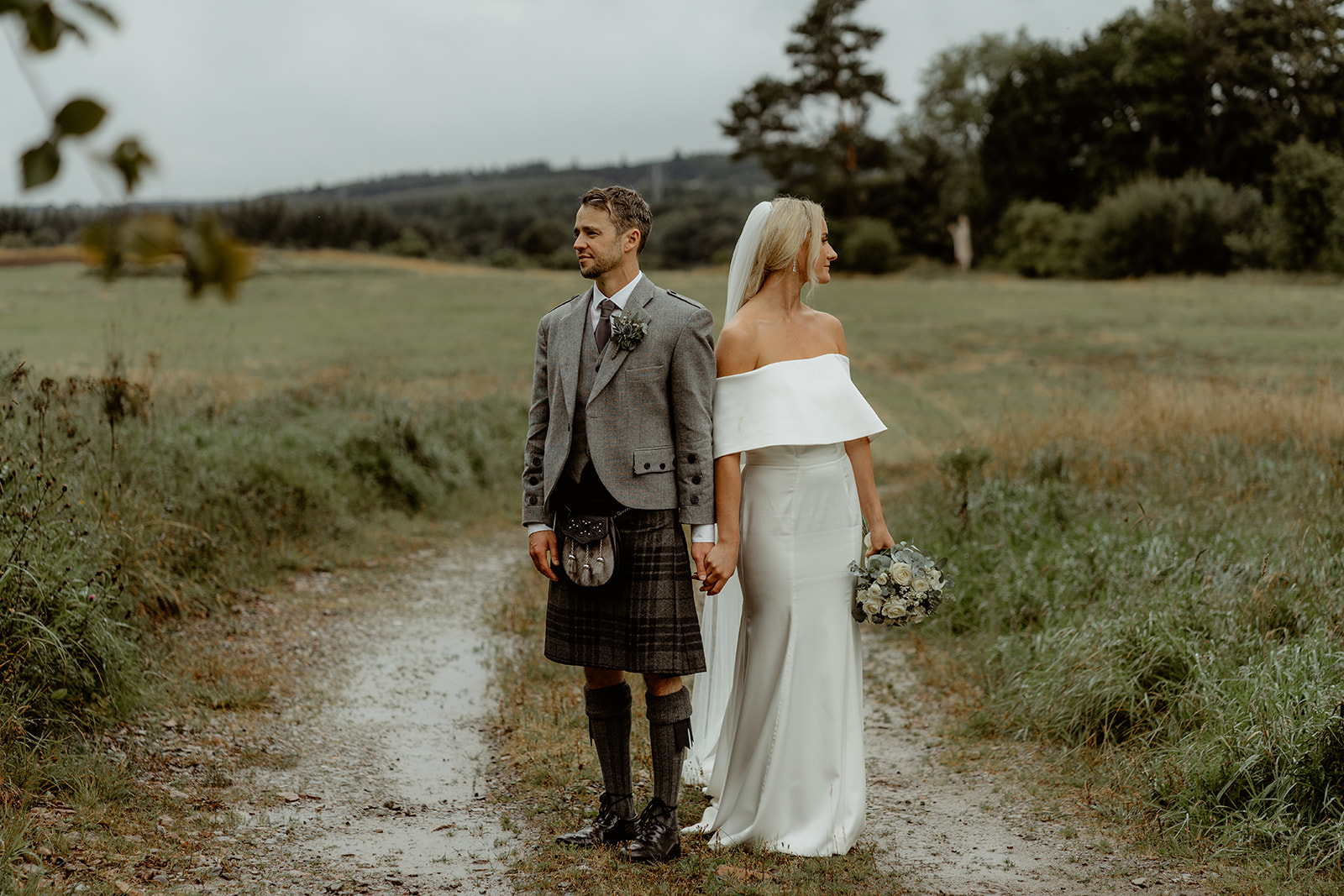 Bride and groom stand side by side holding hands and looking in opposite directions with field in the background
