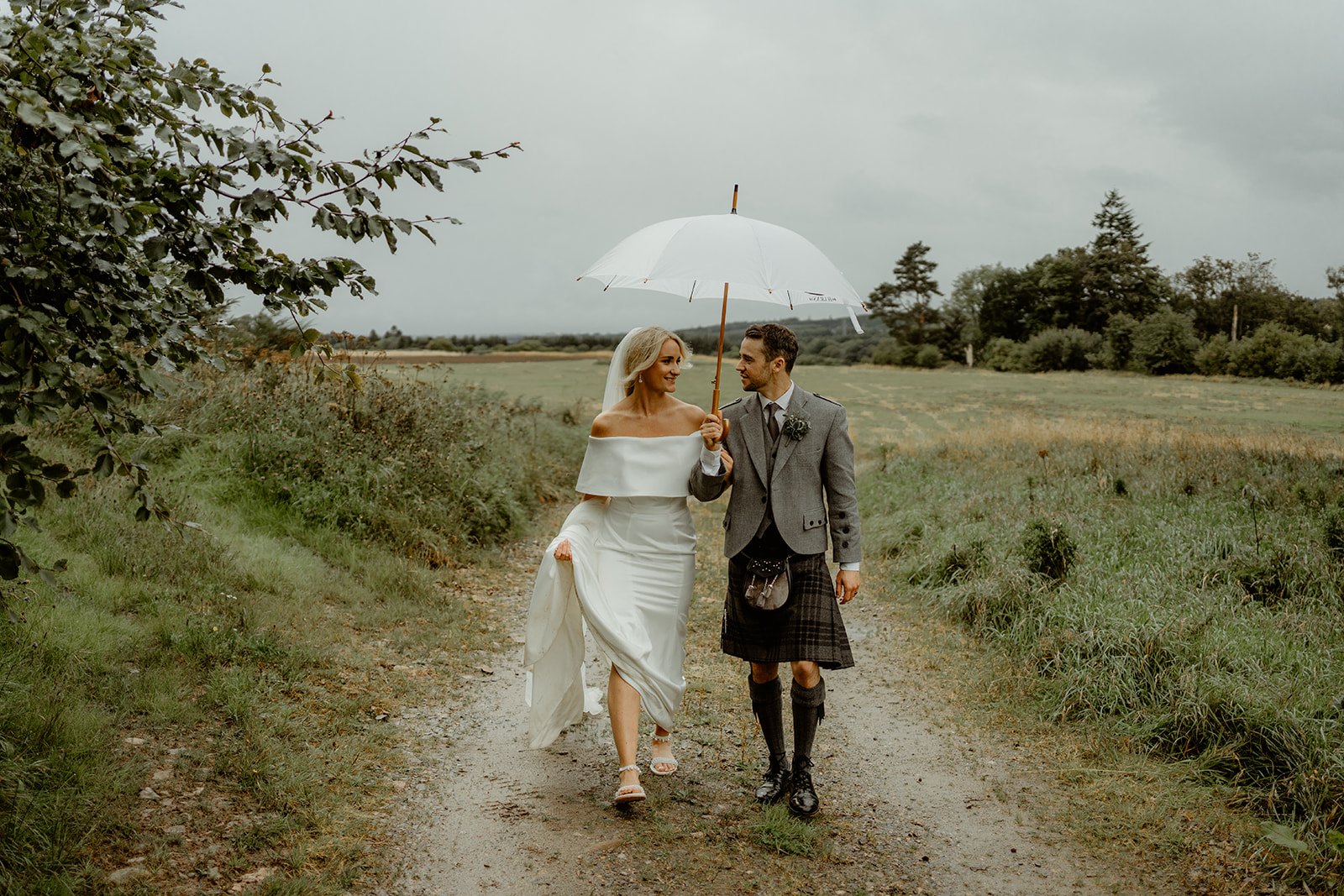 Bride and Groom walk along farm lane holding hands underneath umbrella