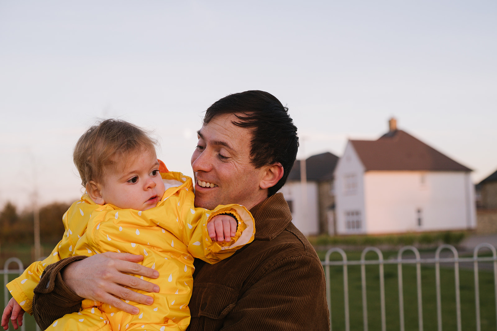 Dad holding his one year old son in the park, its golden hour and pink sunlight is shining on the buildings behind