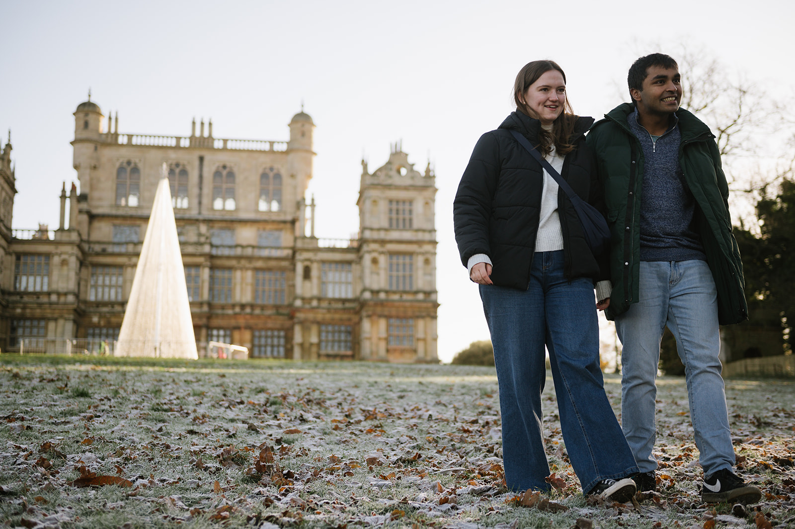 Couple walking with Wollaton Hall and a modern Christmas tree in the background, there's frost on the ground.
