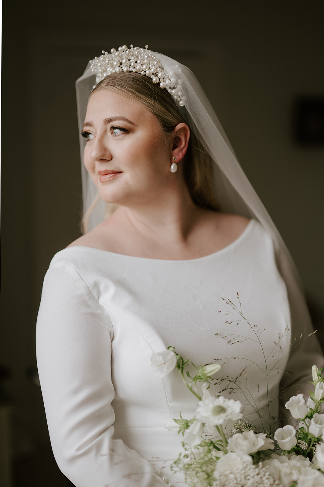bride looking out of window wearing sleeved wedding dress and pearl headband at butley priory