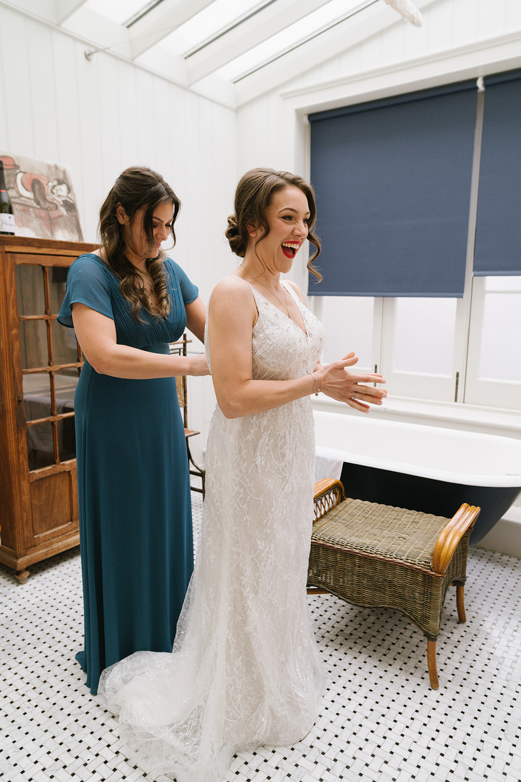 The bride and her sister get the wedding dress on in the light airy bathroom, a roll top bath in the background