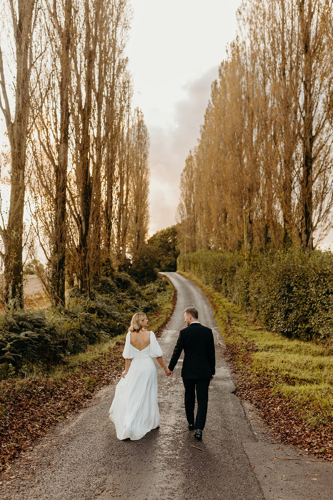 Bride and groom walking together on a pine-lined road leading up to Silchester Farm on their wedding day.