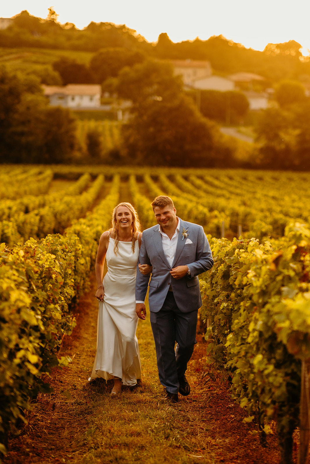 Bride and groom walk down vineyard as the setting sun provides a warm glow