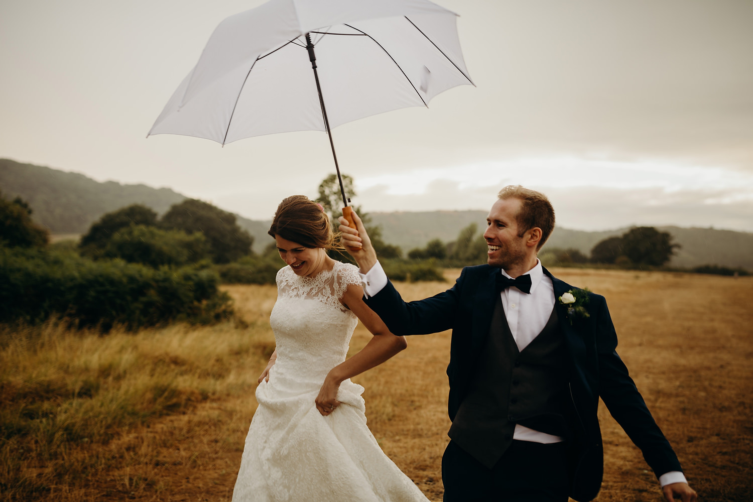 groom holds umbrella as he runs with bride at rainy wedding