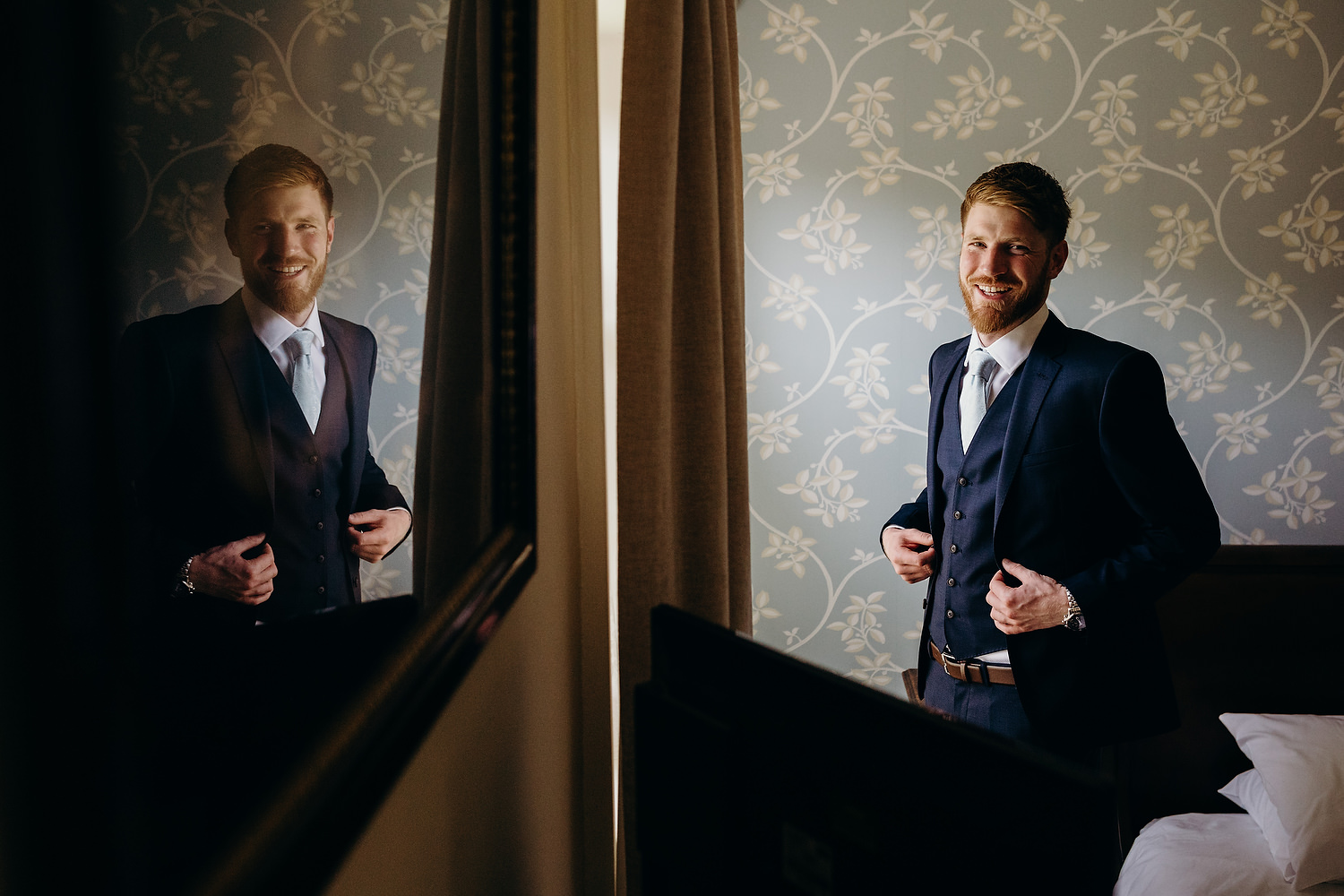 groom gets ready by window with a reflection of him in the mirror