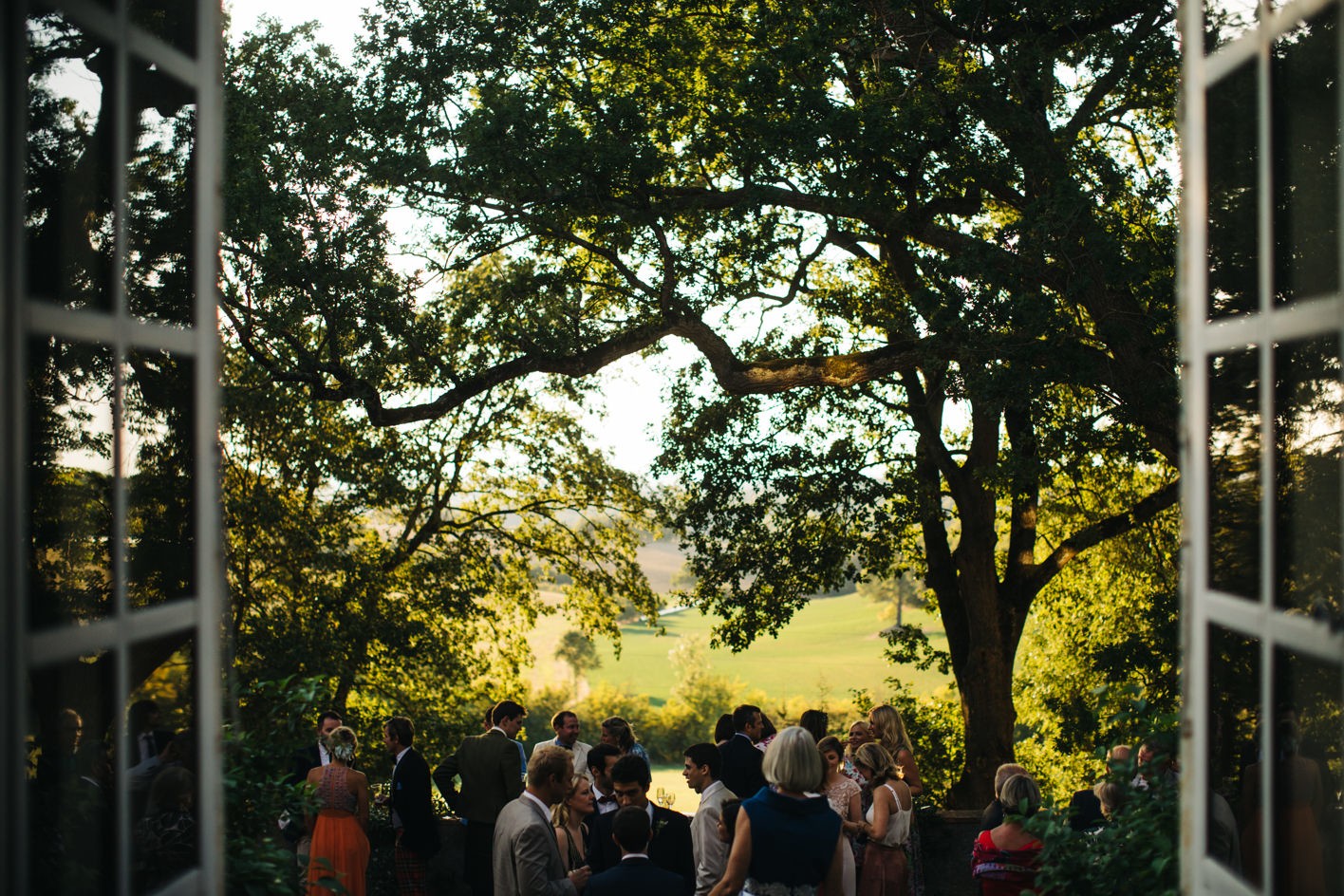 View of guests mingling at wedding drinks reception from window with trees in background