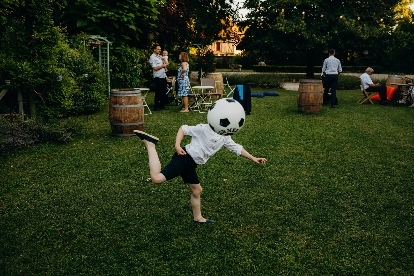Candid image of a child playfully kicking a large soccer ball covering their head in a garden wedding reception